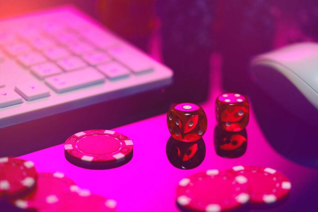 Close-up shot of red dice and red casino chips on a pink surface with a computer keyboard and mouse in the background.