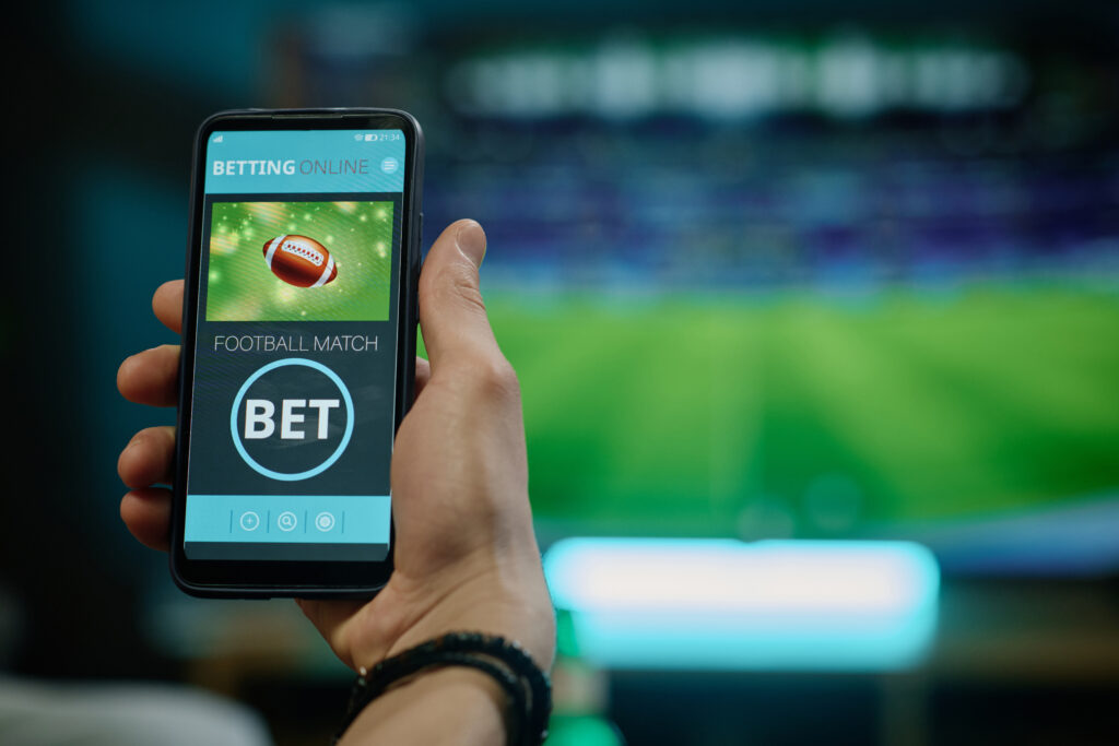 Out of frame man holding a smartphone, with an image of an American football on it. Above this are the words "BETTING ONLINE" and below it are "FOOTBALL MATCH" and "BET" surrounded by a light blue circle. The background of the image is blurred, and in it is a TV showing a stadium field.