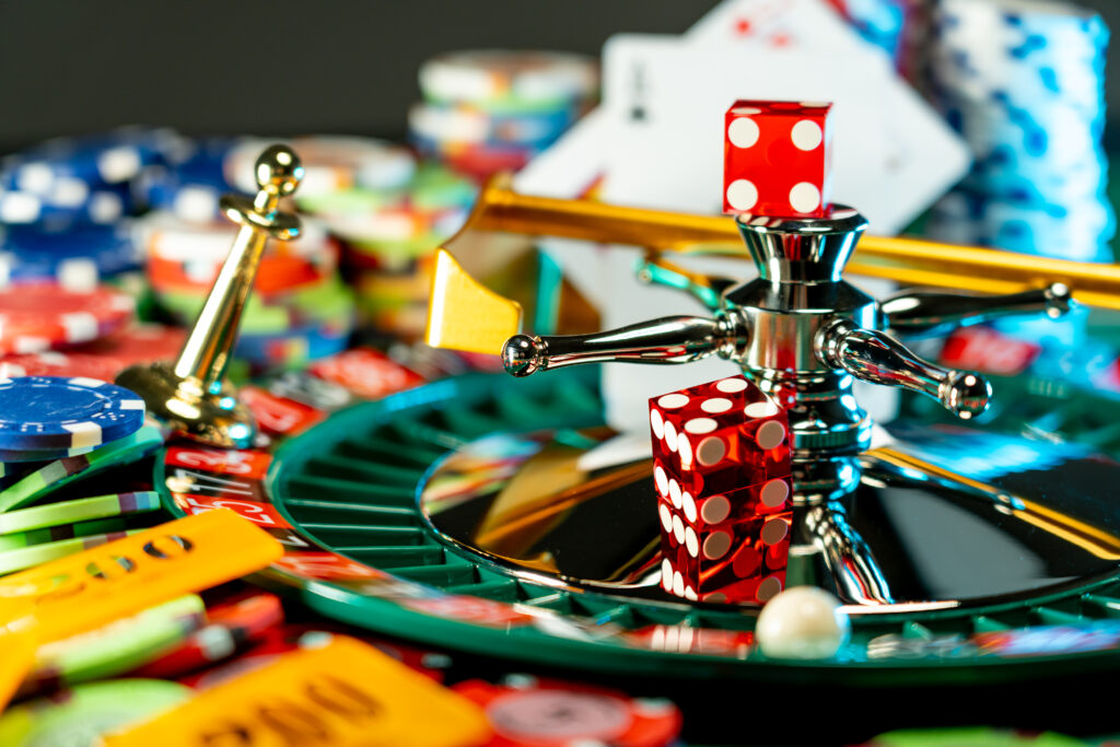 Small roulette wheel with red dice on it. In the background and foreground surrounding it are playing cards and casino chips, blurred.
