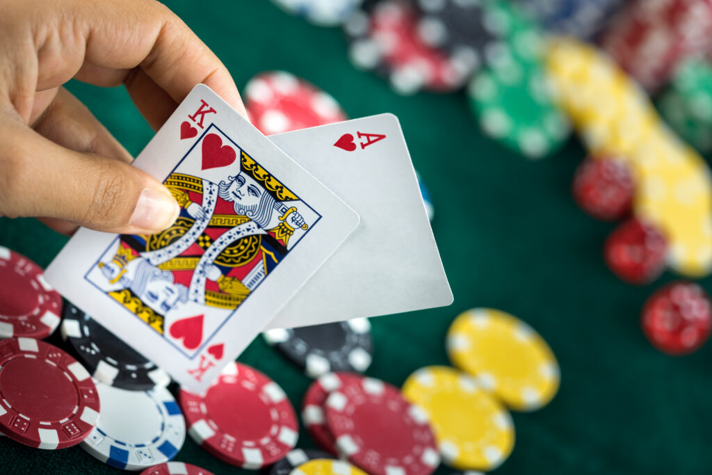Hand holding two playing cards, the King of Hearts and Ace of Hearts, with a blurred green table covered in casino chips in the background.