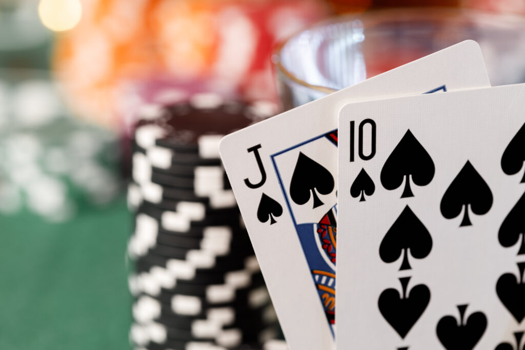 Two playing cards, a Jack of Spades and 10 of Spades, in front of a blurred background of a green casino table, casino chips and and a glass.